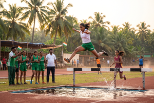 A young female athlete wearing green Goa sports gear clearing a steeplechase water jump hurdle during a track and field competition.