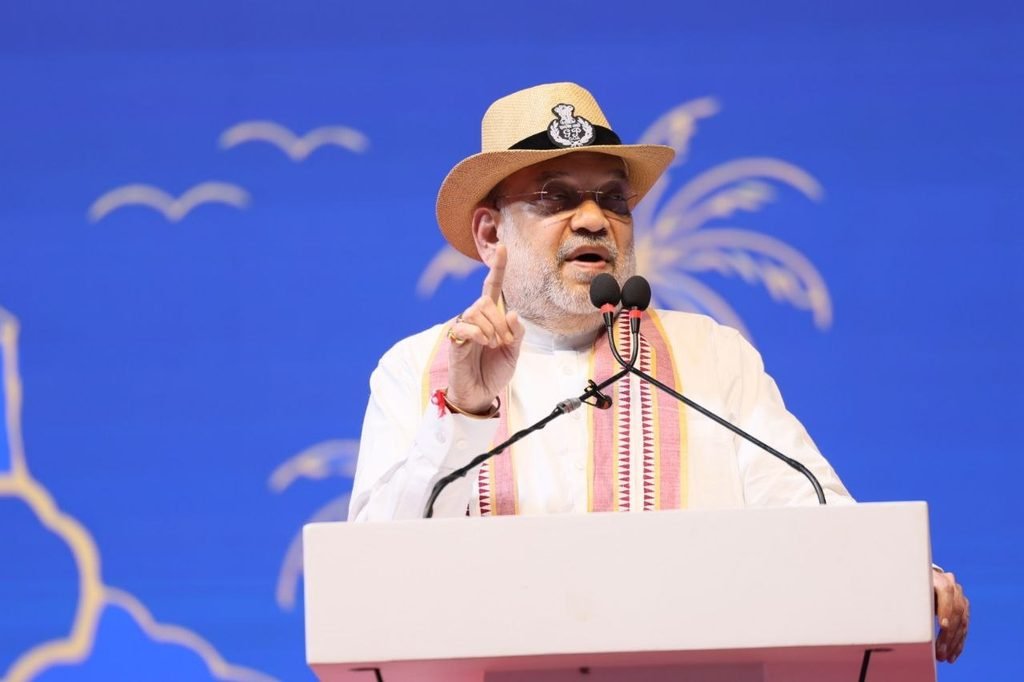 Union Home Minister Amit Shah wearing a hat and traditional shawl while speaking at a podium against a blue backdrop with palm tree graphics.